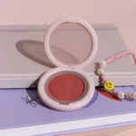 Cuatro unidades de rubor compacto Dolly Blush de MYK Cosmetics en tonos rosa y coral sobre libros decorativos.